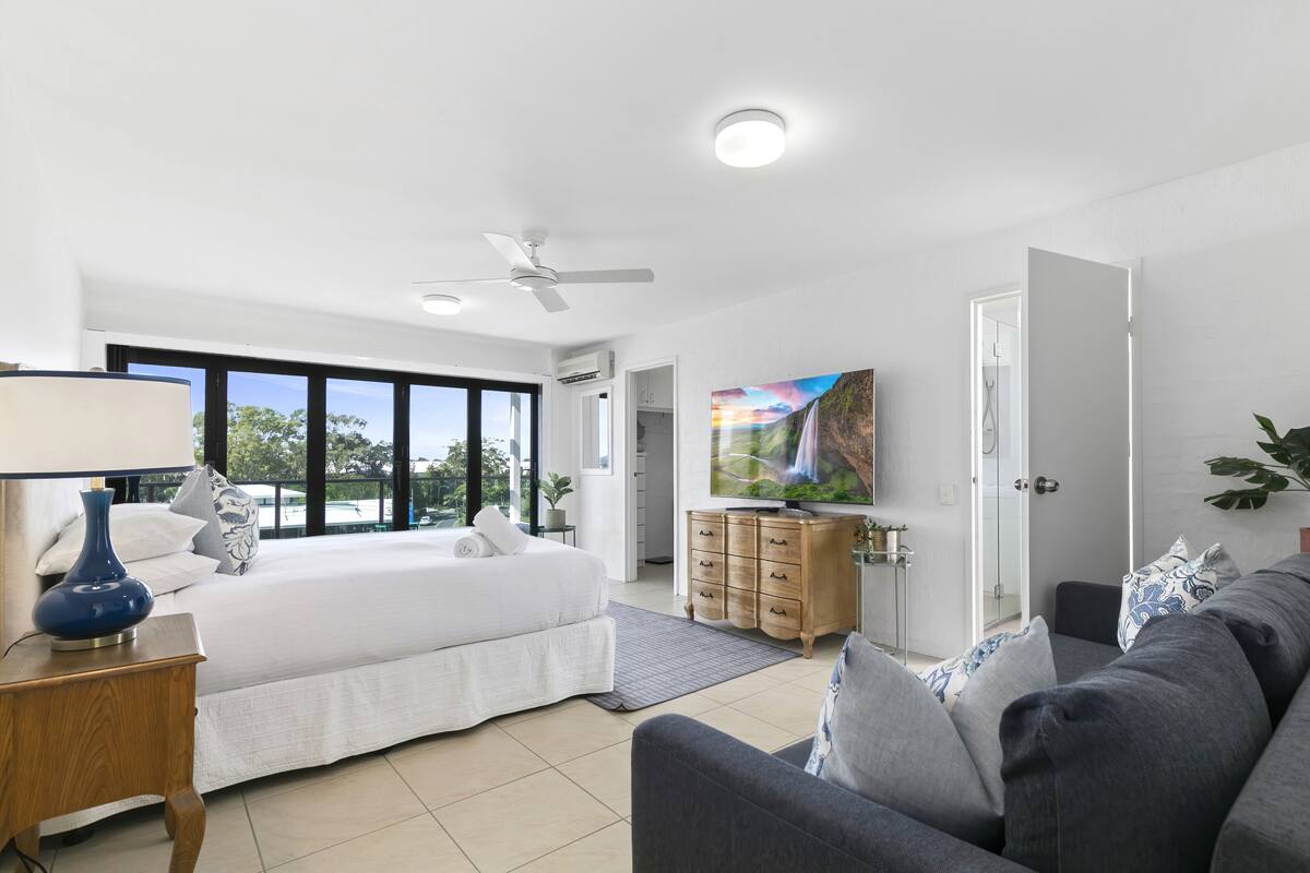 A modern bedroom with a large bed, a TV, a ceiling fan, and a painting.