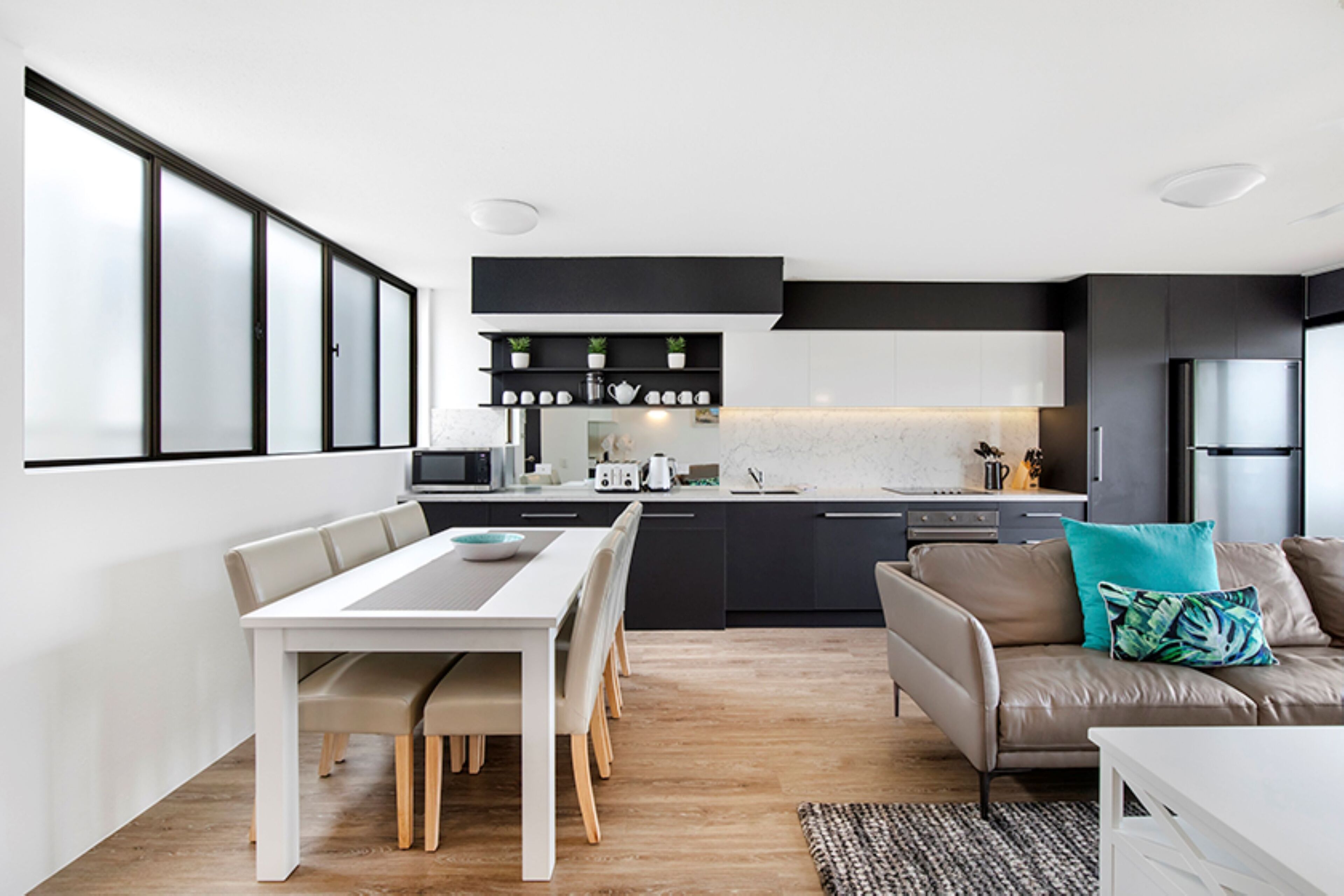 A modern kitchen with dark cabinetry, a dining area with a white table, and a living area with a sofa and cushions.