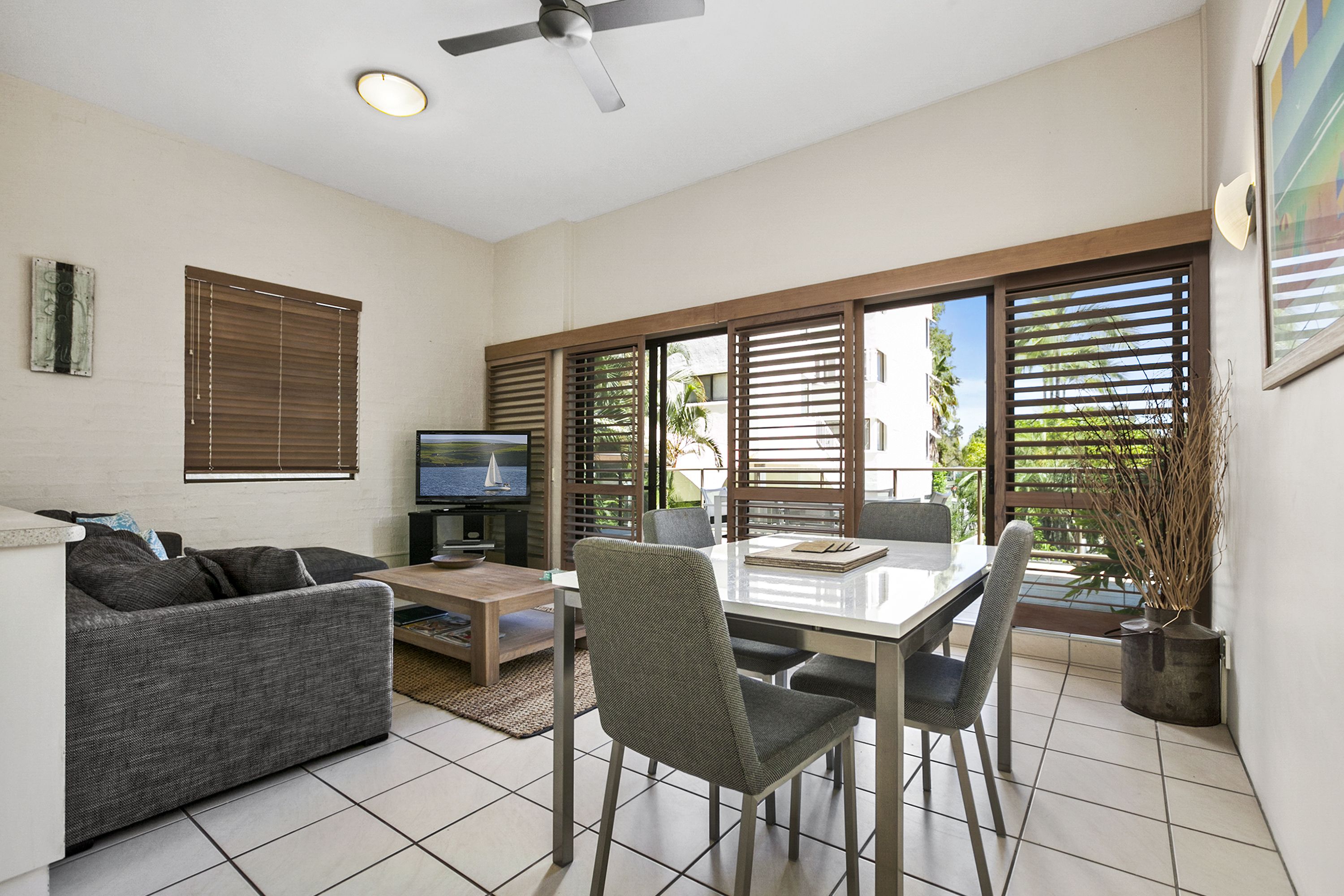 A modern living room with a sofa, dining table, chairs, a TV, and large windows with blinds.
