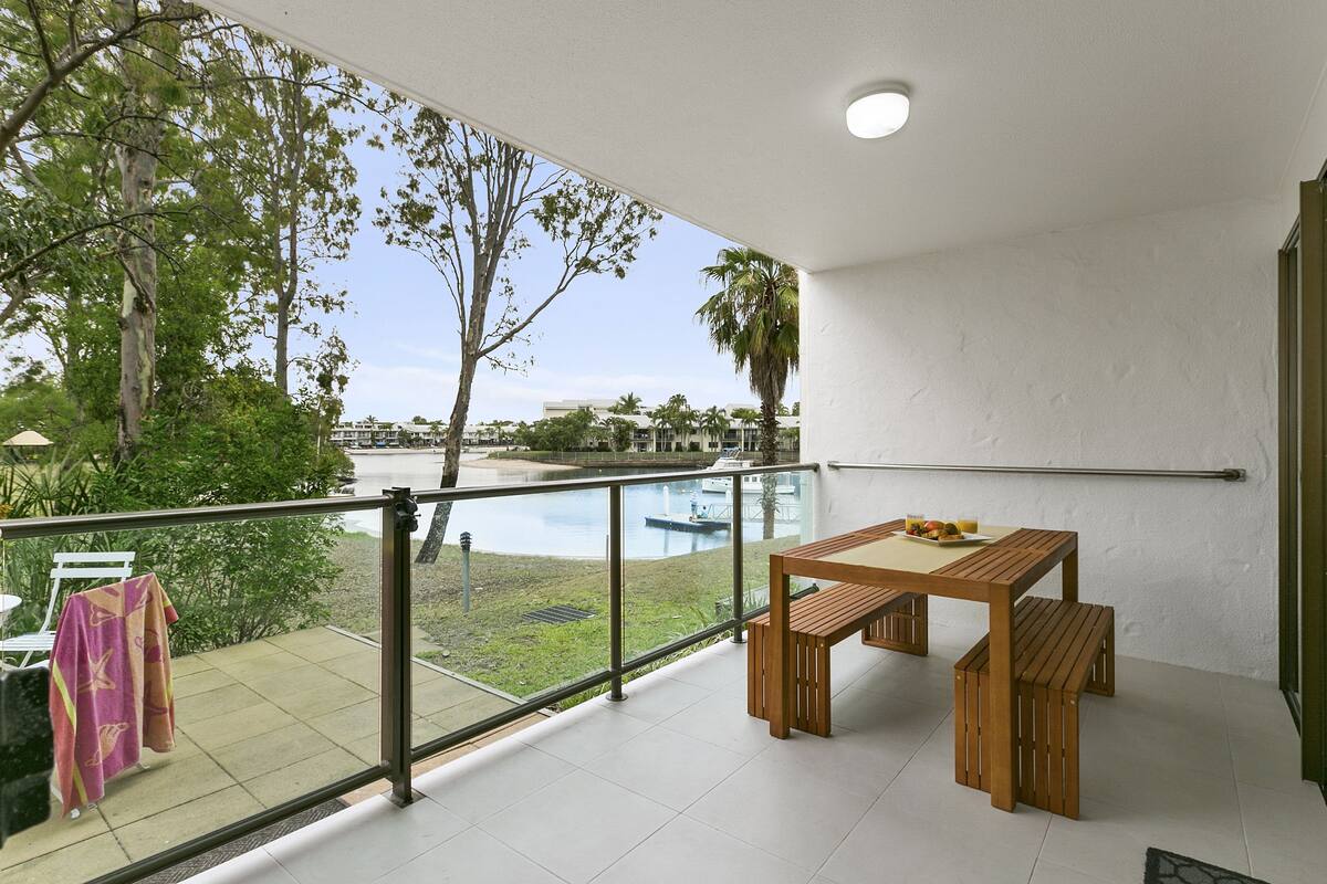 A balcony with a wooden table and bench, a glass railing, and a view of a pool and greenery.