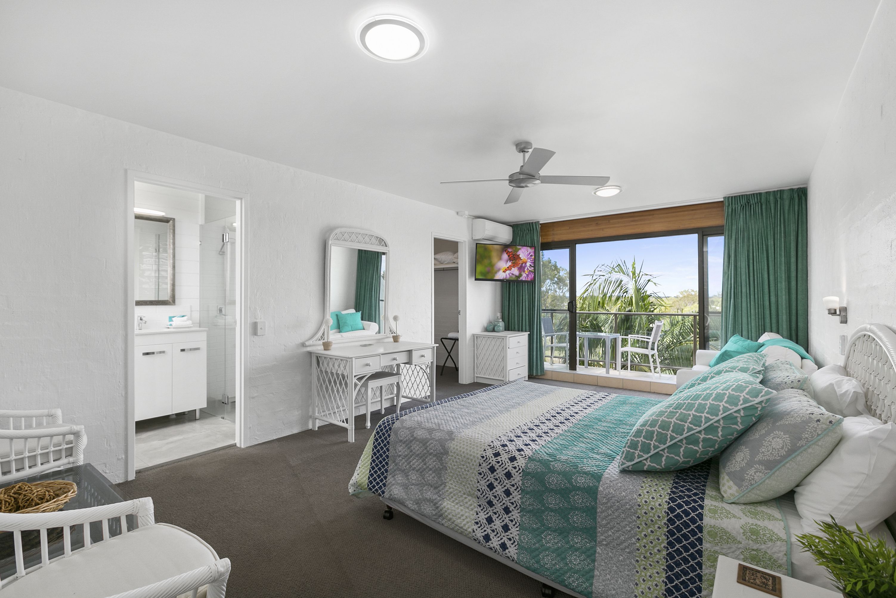 A bedroom with a bed, a desk, a chair, a wardrobe, and a balcony with a view of greenery.