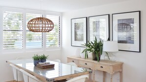 Et moderne spiseområde med et glasbord, hvide stole, et træskænk med en lampe og indrammede billeder på væggen.