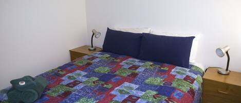 A bed with a colorful quilt, two pillows, a green towel, and two bedside lamps on wooden nightstands.