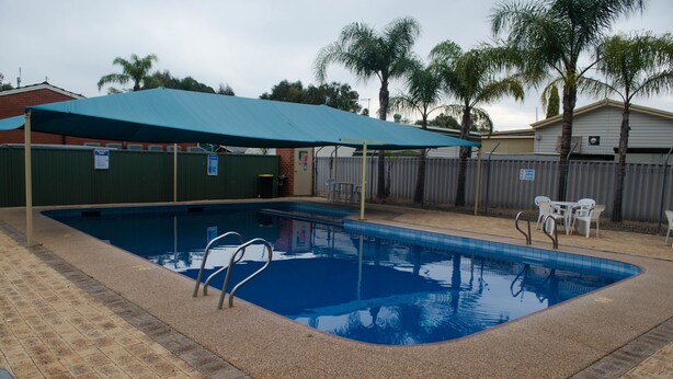 A rectangular swimming pool with a blue tiled interior, surrounded by a brick deck. There are two white chairs and a table under a blue canopy. - Acclaim Swan Valley Tourist Park (West Swan)