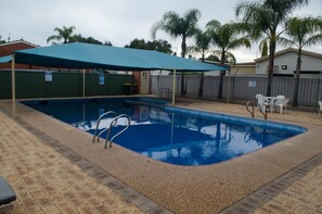 A rectangular swimming pool with a blue tiled interior, surrounded by a brick deck. There are two white chairs and a table under a blue canopy. - Acclaim Swan Valley Tourist Park (West Swan)