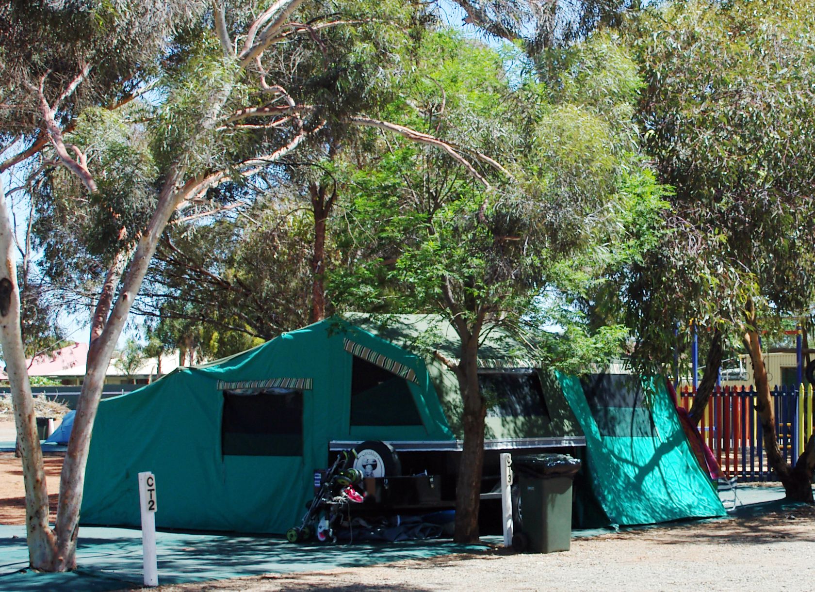 A green tent with a small table and a trash bin nearby, set in a park with trees and a fence in the background.