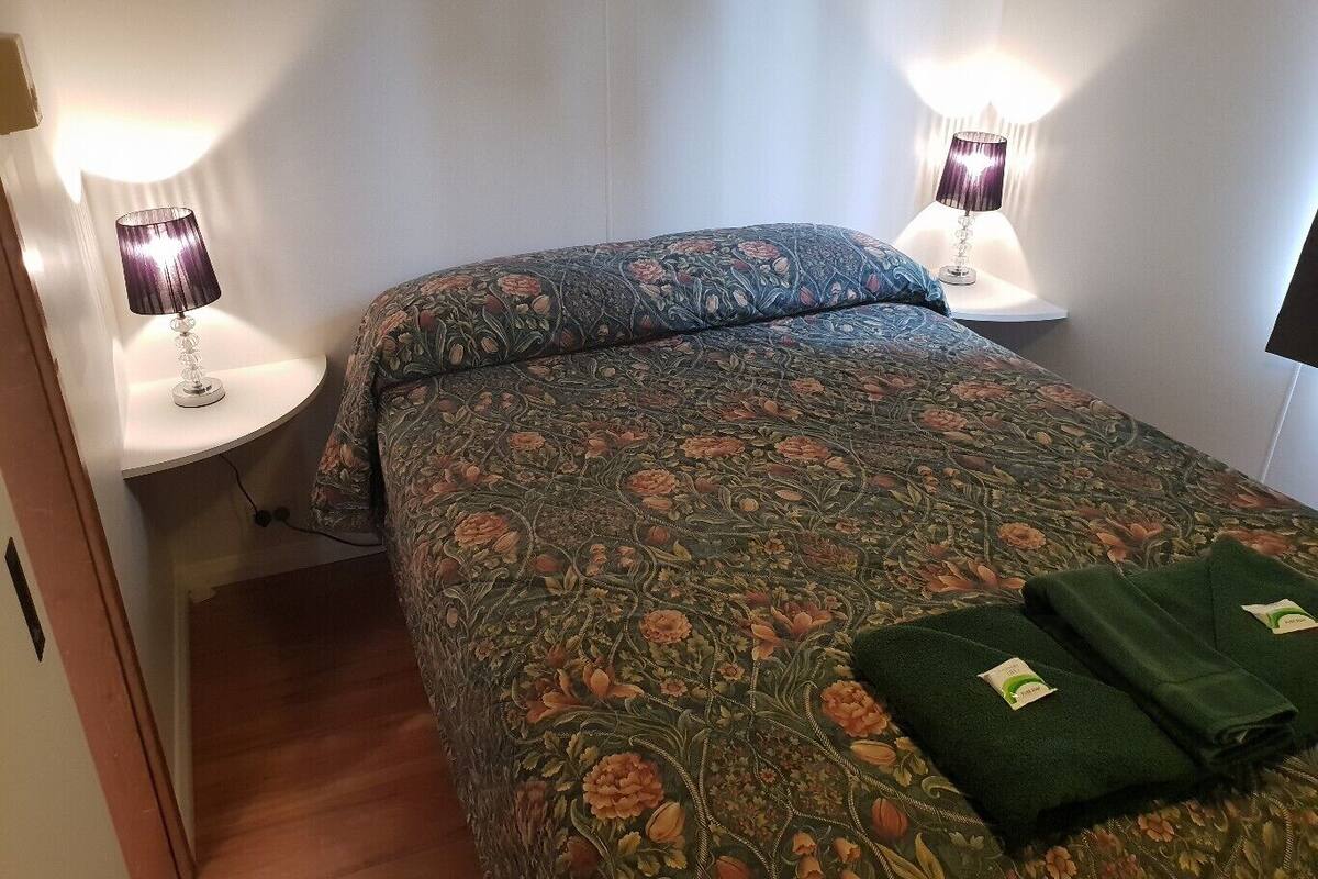 Una cama bien hecha con ropa de cama floral, dos lámparas de noche y toallas dobladas sobre la cama.