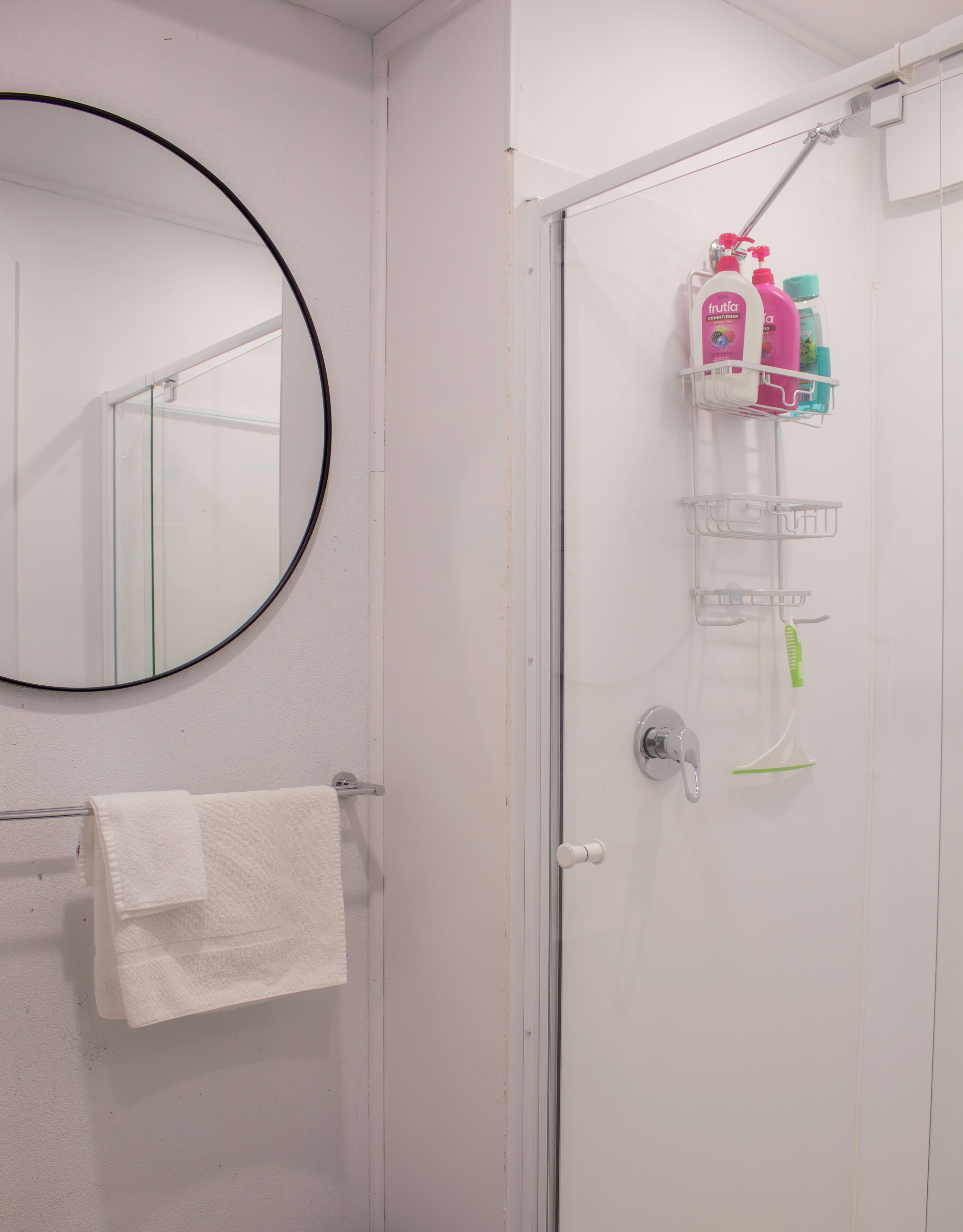 A bathroom with a round mirror, a towel rack with a white towel, a glass shower door, and a shelf with toiletries.