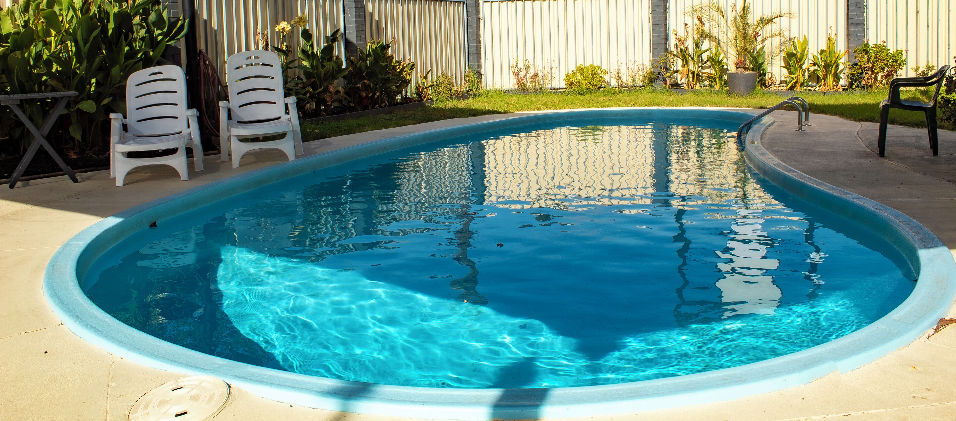 A swimming pool with a curved edge, surrounded by a concrete deck. There are two white plastic chairs and a small table on the deck.