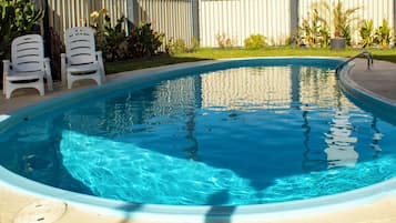 A swimming pool with a curved edge, surrounded by a concrete deck. There are two white plastic chairs and a small table on the deck.
