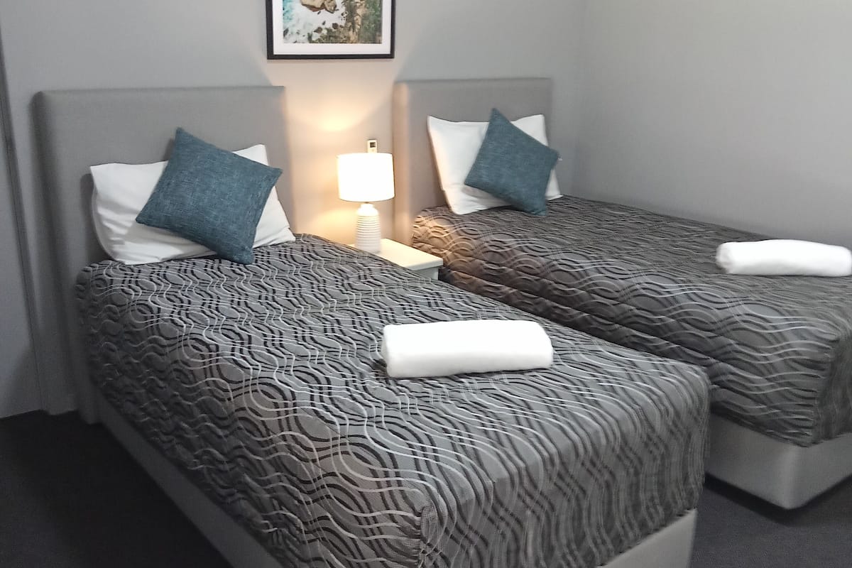Quarto de hotel com duas camas, cada uma com roupa de cama e almofadas estampadas, uma mesa de cabeceira com abajur e um quadro na parede.