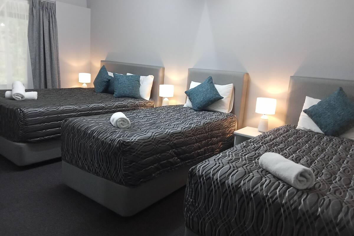 Quarto de hotel com três camas, cada uma com colcha estampada e almofadas, e luminárias laterais.
