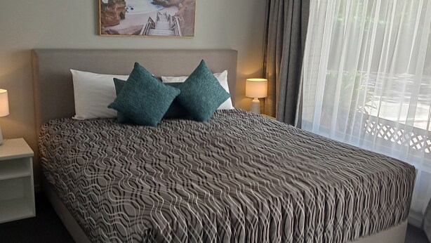 A large bed with a patterned bedspread, two pillows, a nightstand with a lamp, a painting on the wall, and a window with curtains.