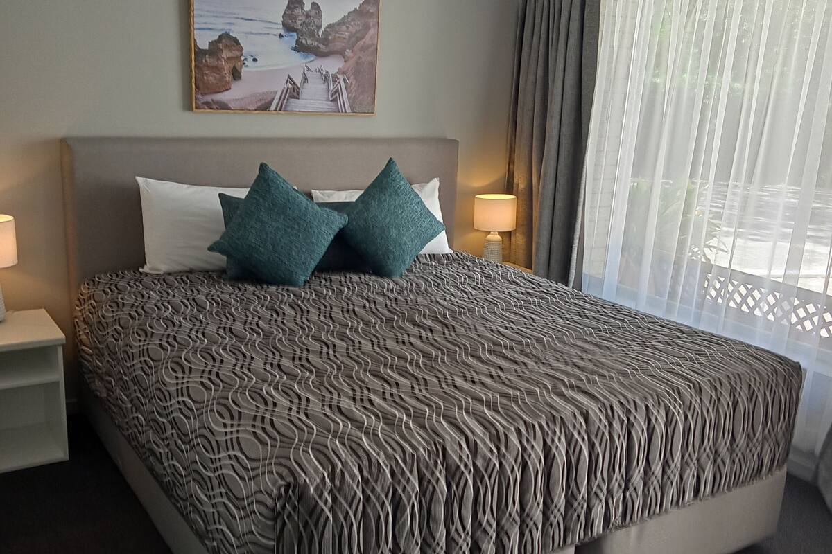 Uma cama grande com colcha estampada, duas almofadas, mesa de cabeceira com abajur, quadro na parede e janela com cortinas.