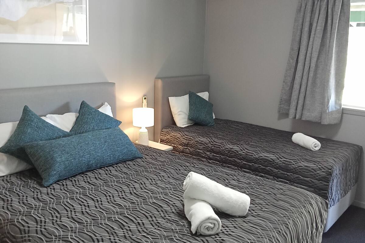 Quarto de hotel com duas camas, paredes cinzas, um quadro, um ar condicionado e uma janela com cortinas.