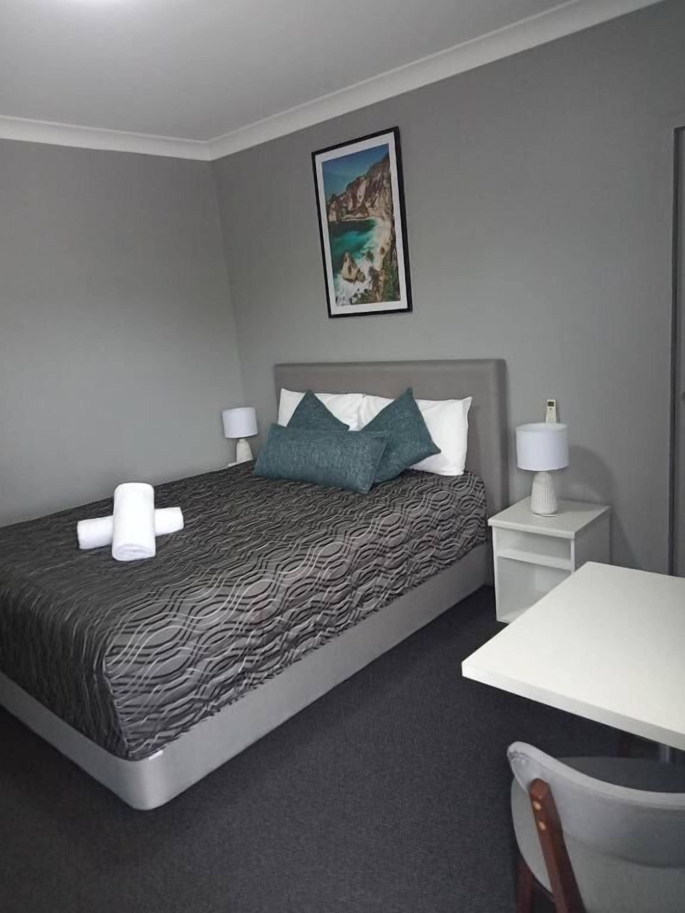A hotel room with a bed, a bedside table, a chair, and a painting on the wall.