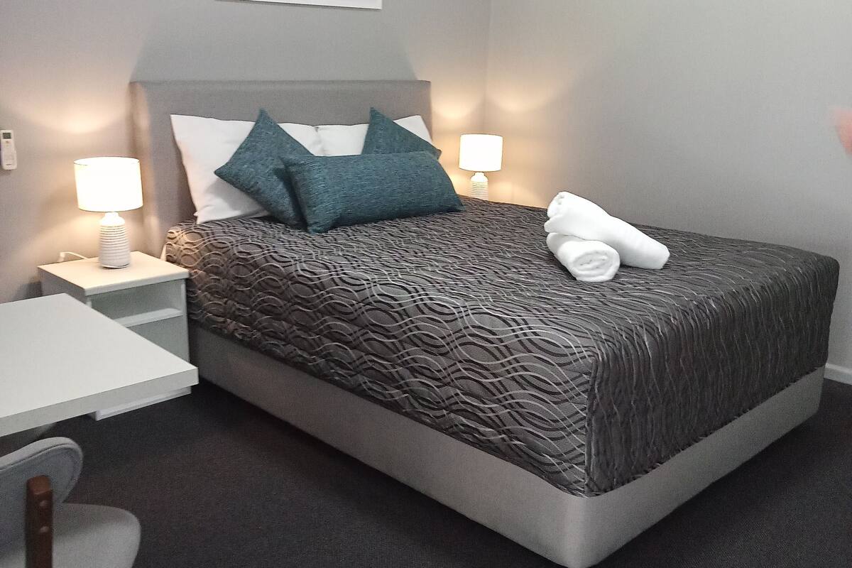 Cama bem arrumada com colcha estampada, duas almofadas e uma toalha enrolada. Há uma pequena mesa de cabeceira com uma luminária, uma cadeira e uma obra de arte pendurada na parede acima da cama.