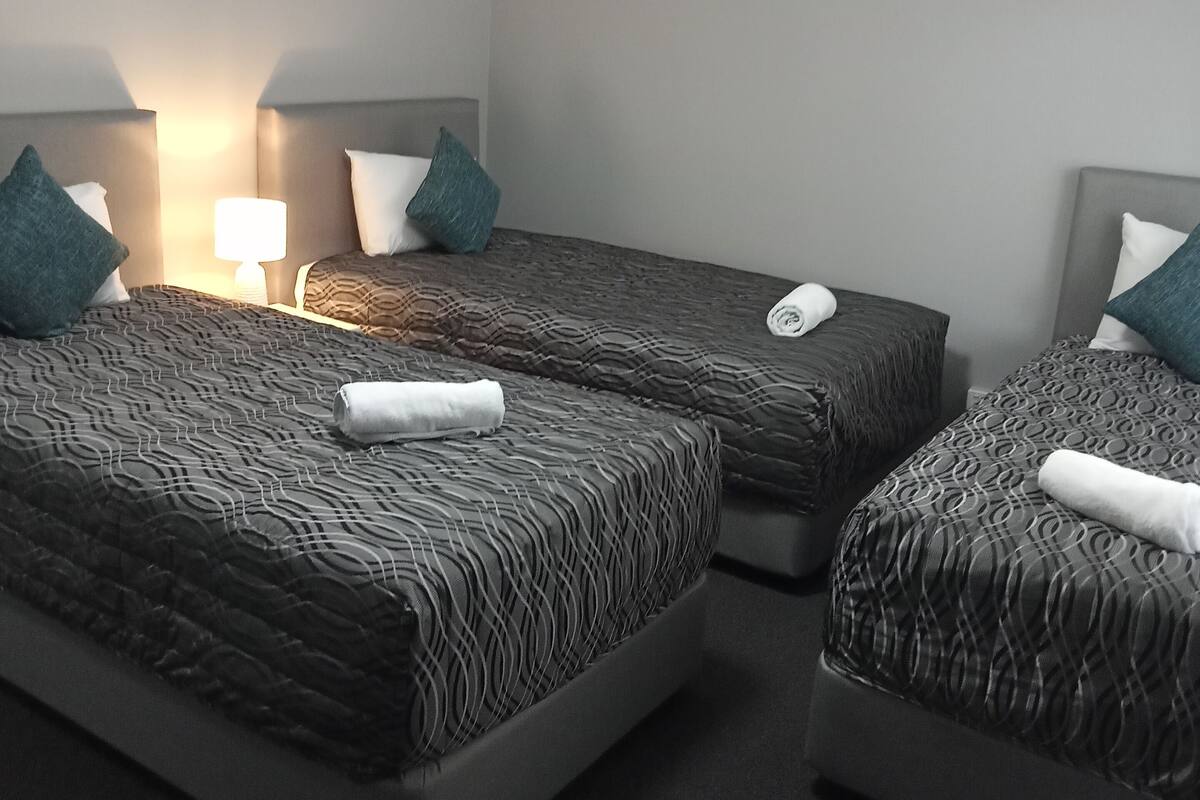 Quarto de hotel com duas camas, cada uma com cabeceira, luminária de cabeceira e uma toalha enrolada sobre a cama.