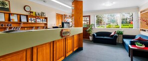 A reception area with a wooden desk, a reception sign, a clock, and a window with a view of greenery. - Solomon Inn Wollongong (Figtree)