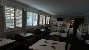 A restaurant with multiple tables and chairs, large windows with blinds, and a counter area with a display case.