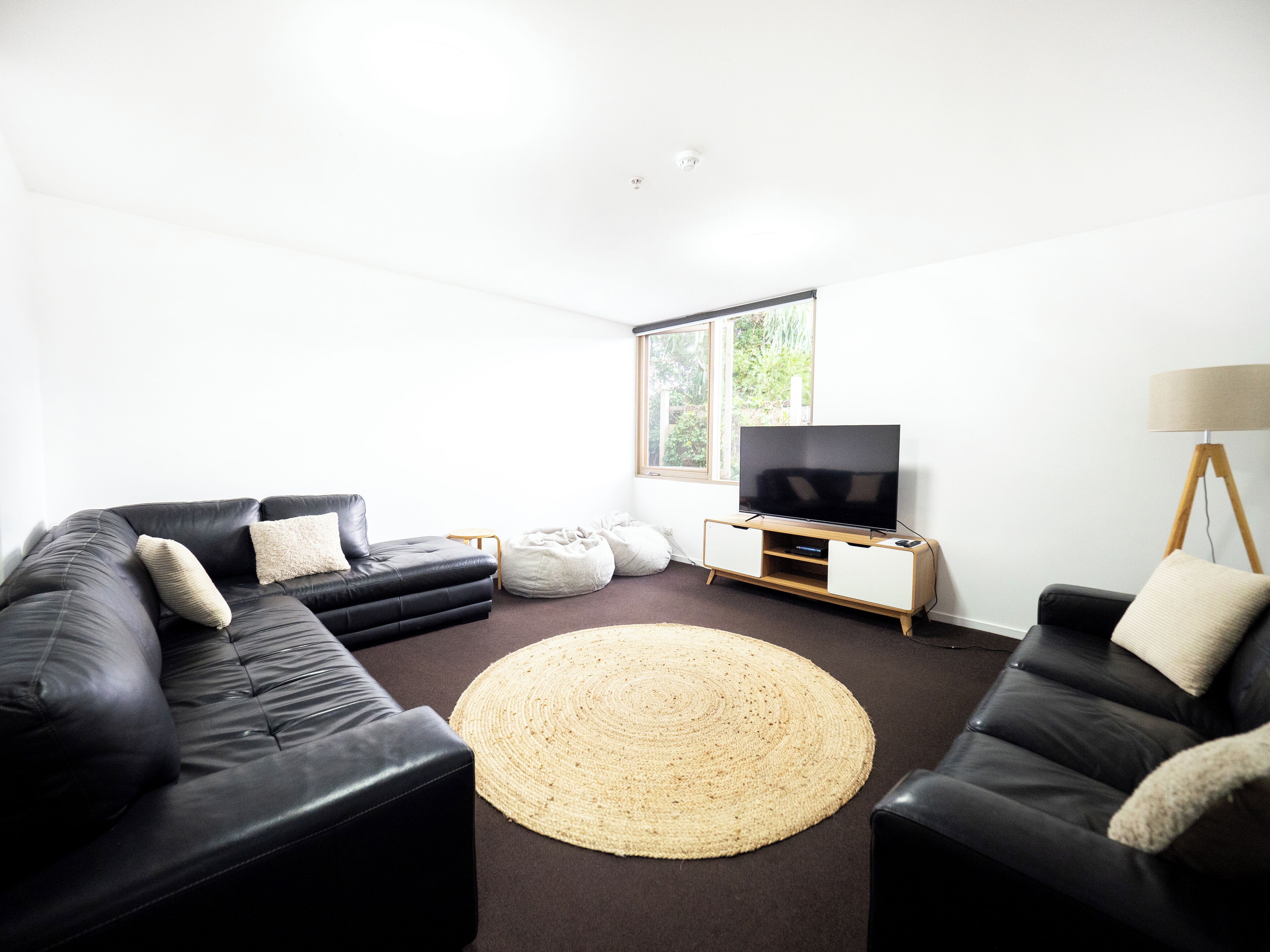 Ein modernes Wohnzimmer mit zwei schwarzen Ledersofas, einem großen Flachbildfernseher auf einem Holzbodenständer, einem runden Teppich aus Gewebe und einer Stehlampe.