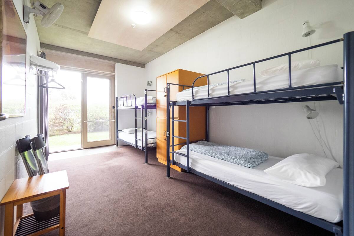 A dormitory room with bunk beds, a wooden desk, and a sliding glass door leading to an outdoor area.