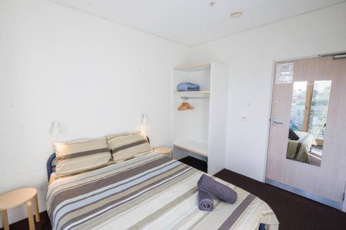 A single bed with a striped bedspread, a bedside table, a wardrobe, and a door leading outside.