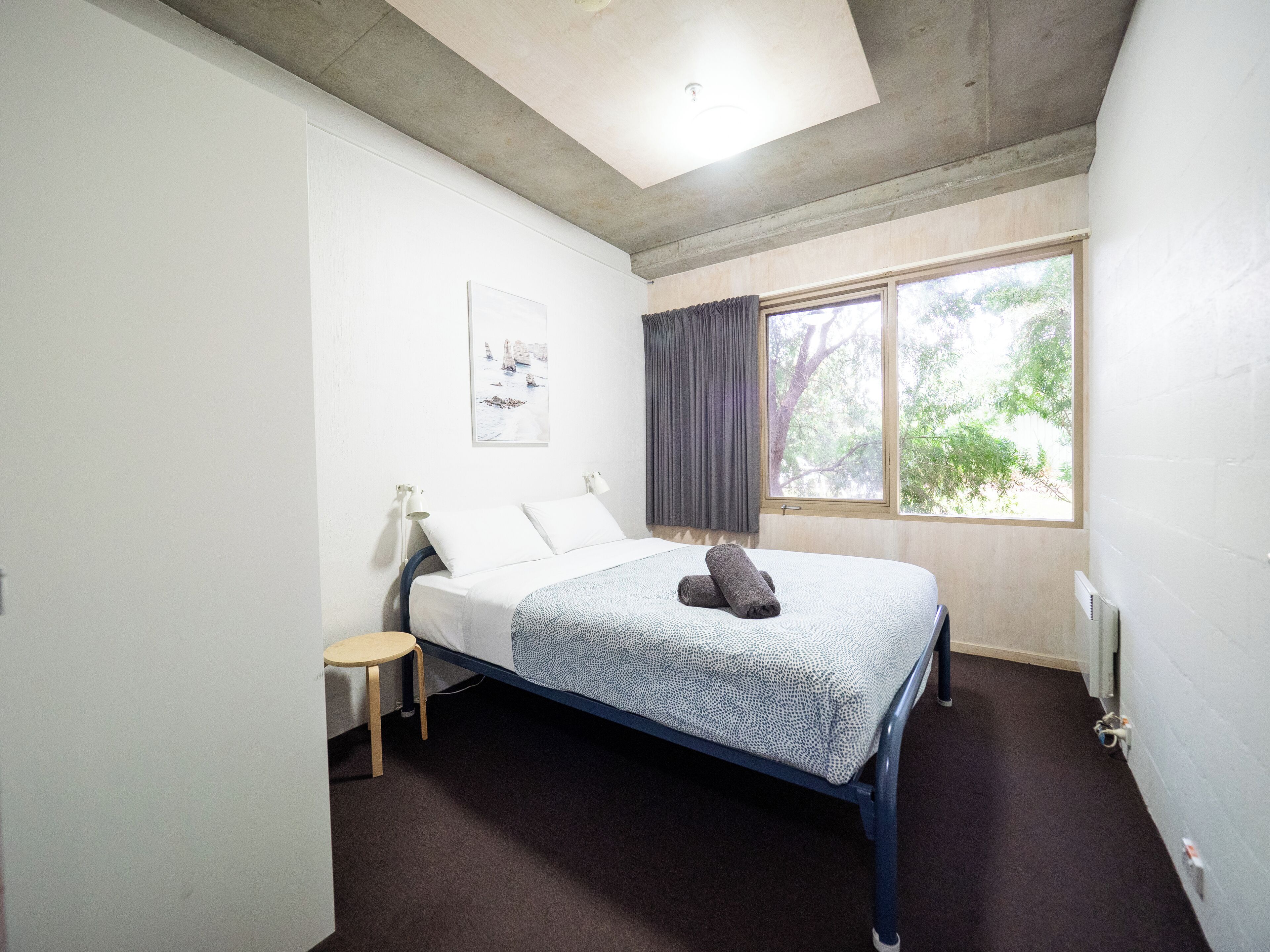 Ein Schlafzimmer mit Bett, kleinem Tisch, Fenster mit Vorhängen und Deckenleuchte.