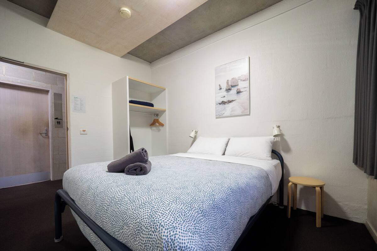 A neatly made bed with a patterned bedspread, a bedside table with a lamp, a small stool, and a painting on the wall.