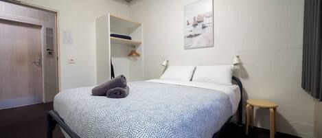 A neatly made bed with a patterned bedspread, a bedside table with a lamp, a small stool, and a painting on the wall.