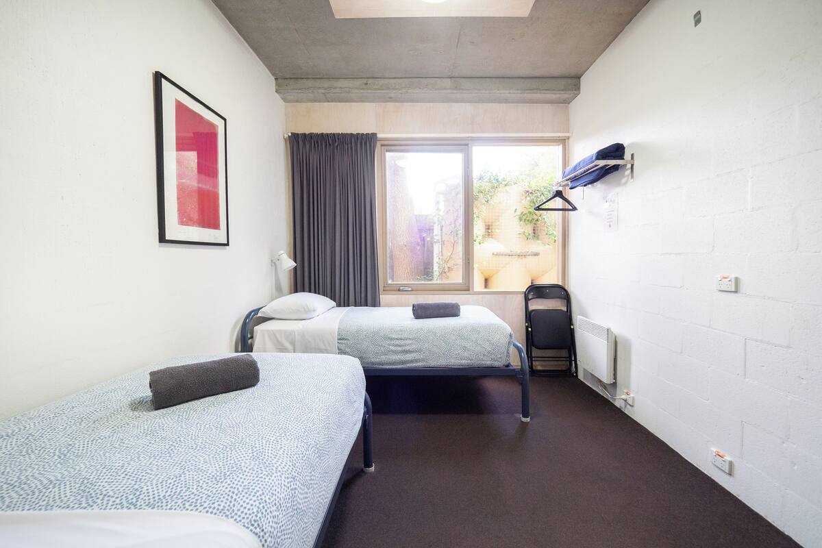 A room with two beds, a window with curtains, a radiator, and a wall-mounted shelf with clothes.