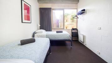 A room with two beds, a window with curtains, a radiator, and a wall-mounted shelf with clothes.