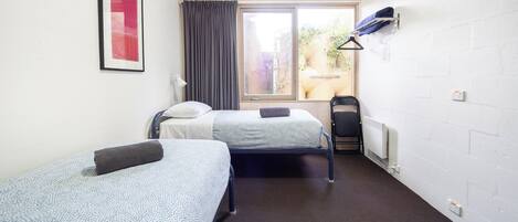A room with two beds, a window with curtains, a radiator, and a wall-mounted shelf with clothes.