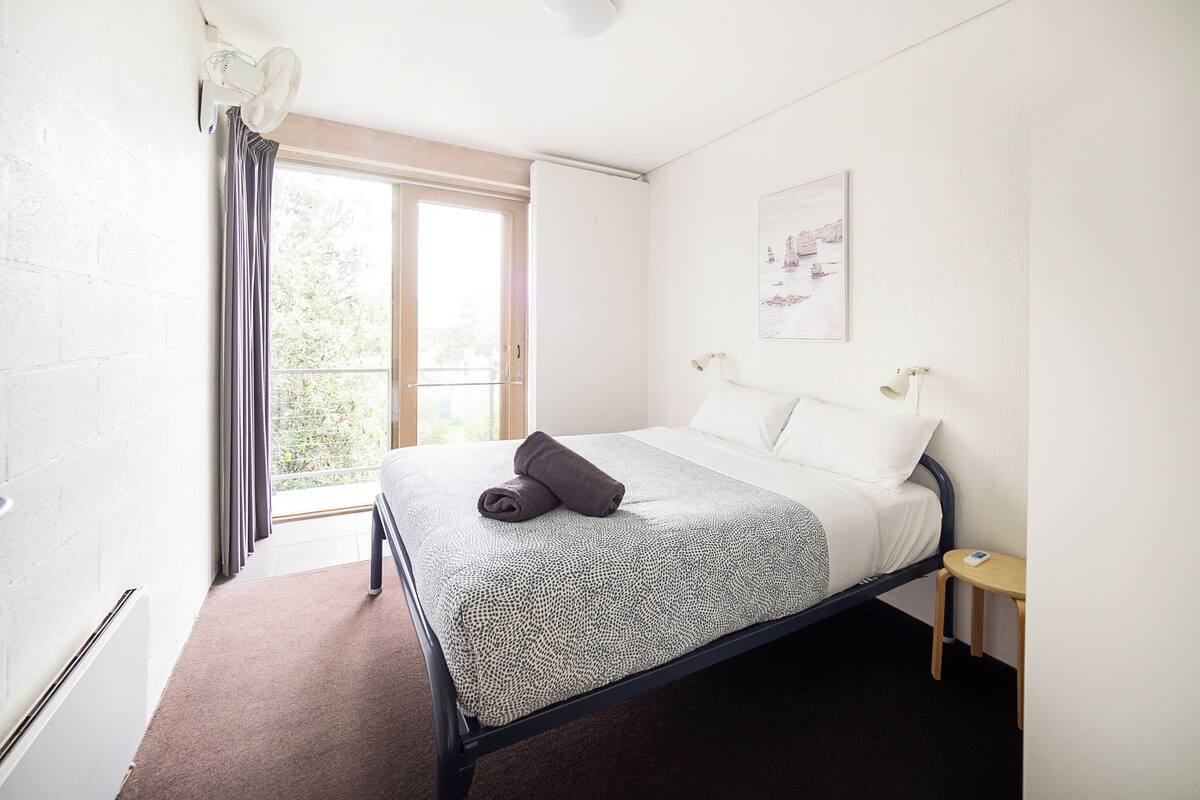 A bedroom with a bed, a bedside table, a window with curtains, and a wall-mounted light fixture.
