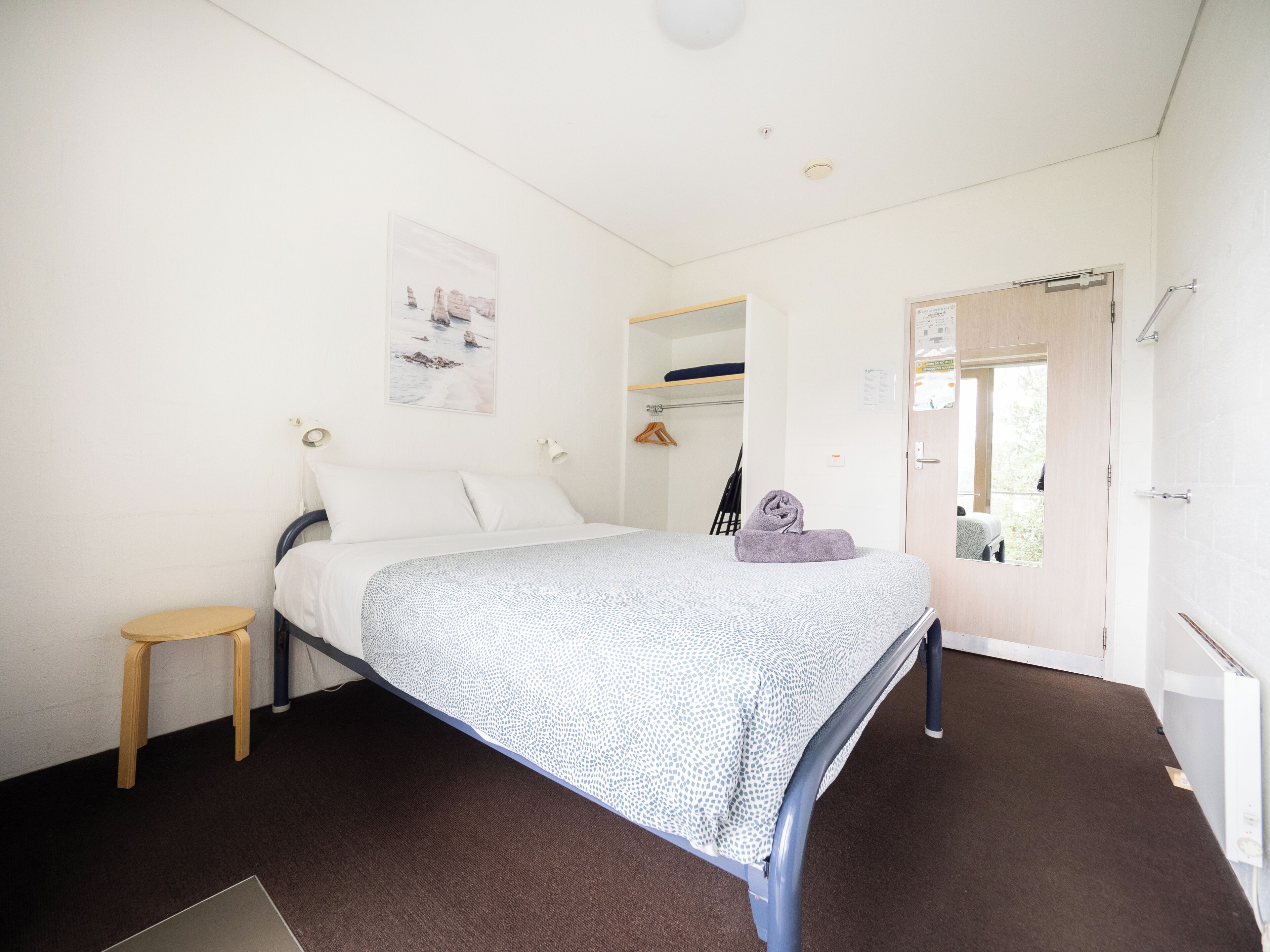 Una habitación con cama individual, una colcha estampada, un taburete de madera, un cuadro en la pared y una puerta que da a un baño.