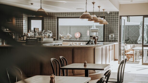 Un restaurante con mesas y sillas de madera, una zona de bar con botellas, y un amplio ventanal al fondo.