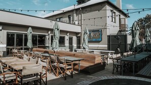 Un patio al aire libre con mesas y sillas de madera, un hogar de fuego de ladrillo y luces colgantes.