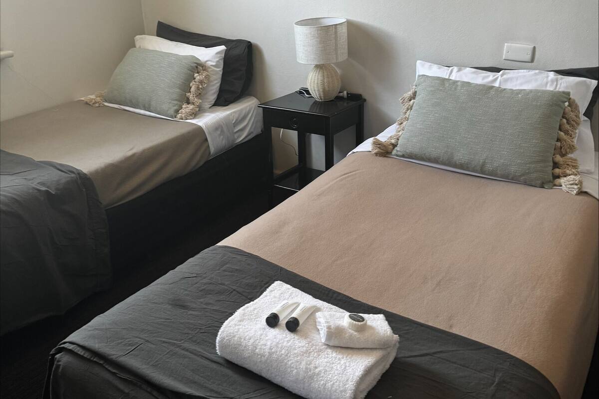 Habitación de hotel con dos camas, cada una con una almohada y una manta. Entre las camas, una mesita de noche con una lámpara.