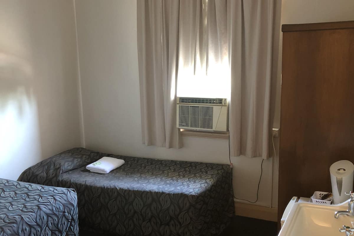 Una habitación con dos camas, una ventana con cortinas, un aire acondicionado, un lavamanos y un cubo de basura.