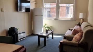 A compact living space with a kitchenette, a sofa, a small table, and a window with blinds.