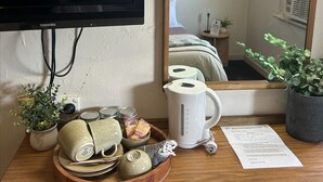Habitación de hotel con televisión de pantalla plana montada en la pared, una mesita de madera con un juego de té y una tetera blanca, además de una planta en maceta.