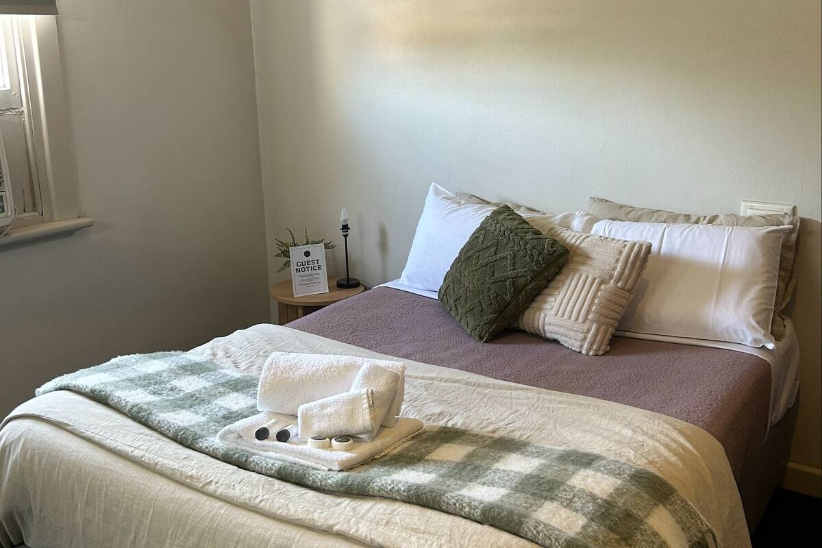 Una cama bien hecha con una colcha a cuadros, un almohadón verde y un cartel que indica un aviso para huéspedes.