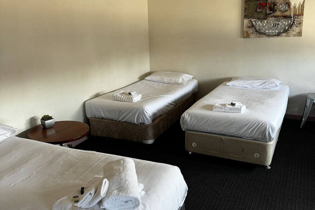 Habitación de hotel con tres camas, una pequeña mesa redonda con una planta y una obra de arte colgada en la pared.