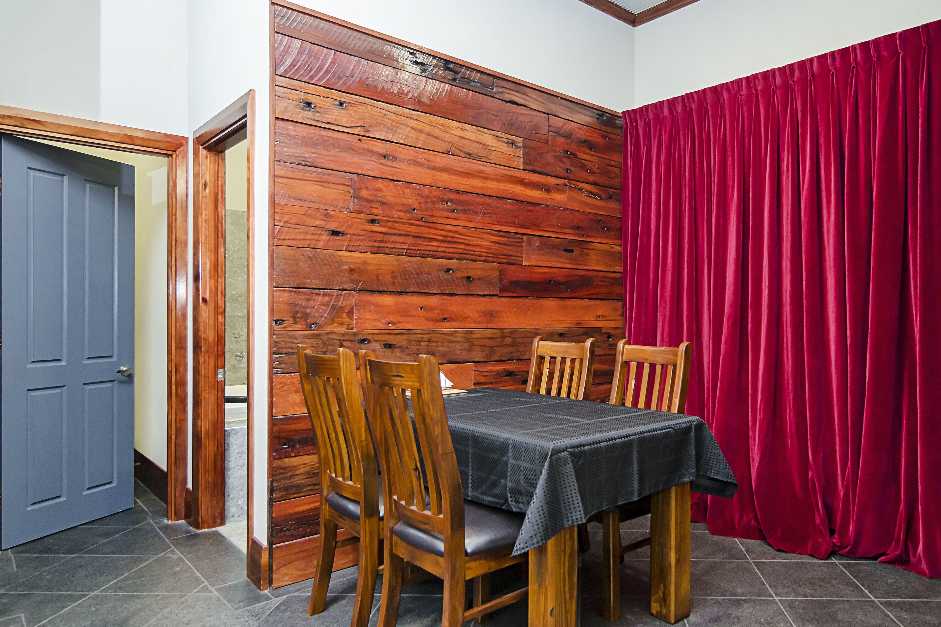 A dining area with a wooden wall, a table with a tablecloth, wooden chairs, and a red curtain.