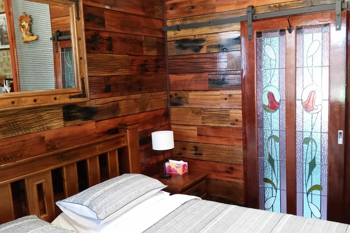 Quarto de madeira com cama, mesa de cabeceira, abajur e porta com vitrais.
