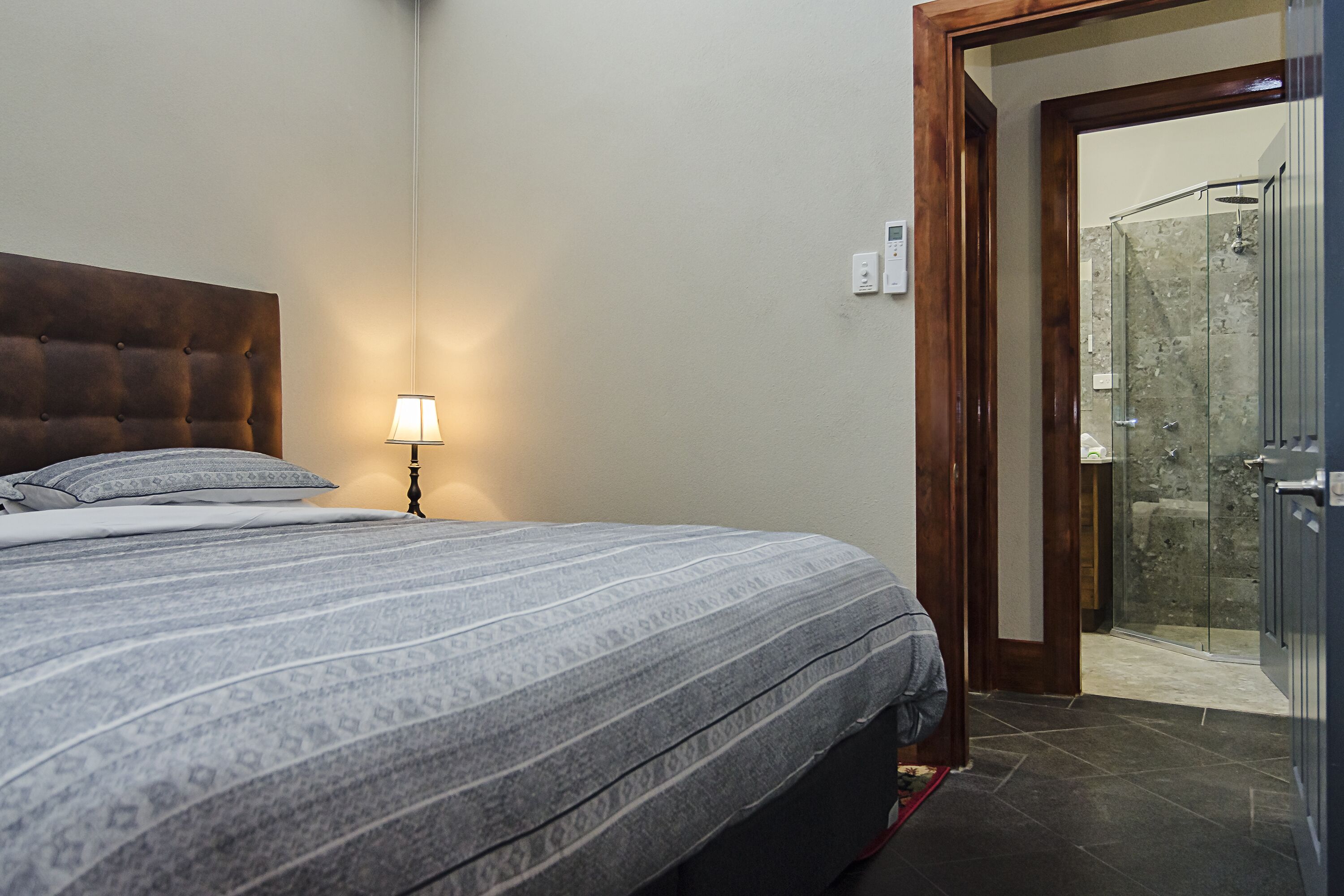 A bedroom with a large bed, a bedside lamp, and a door leading to a bathroom with a shower.
