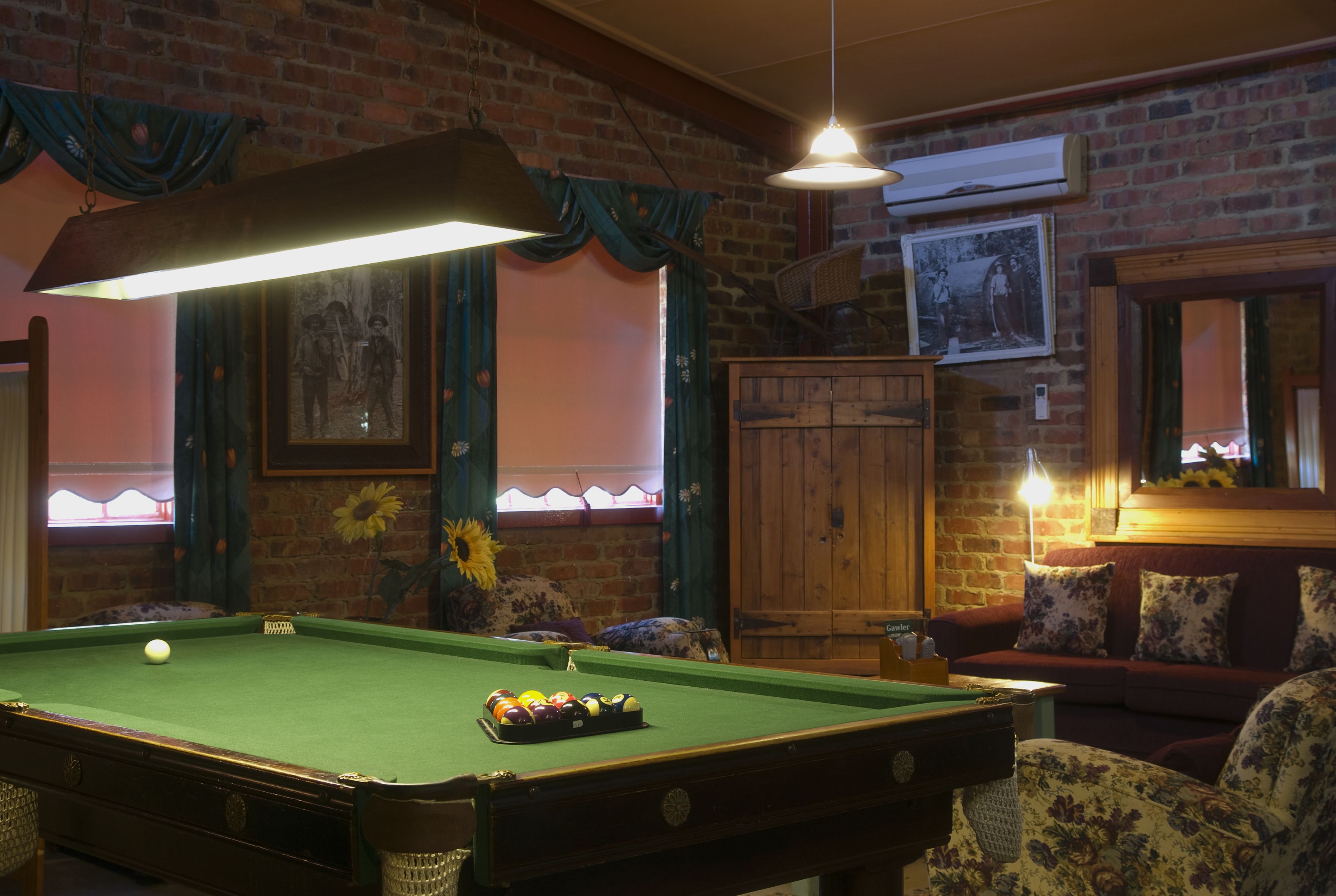 A pool table with balls, a sofa, and a wooden cabinet in a room with brick walls and a hanging light fixture.