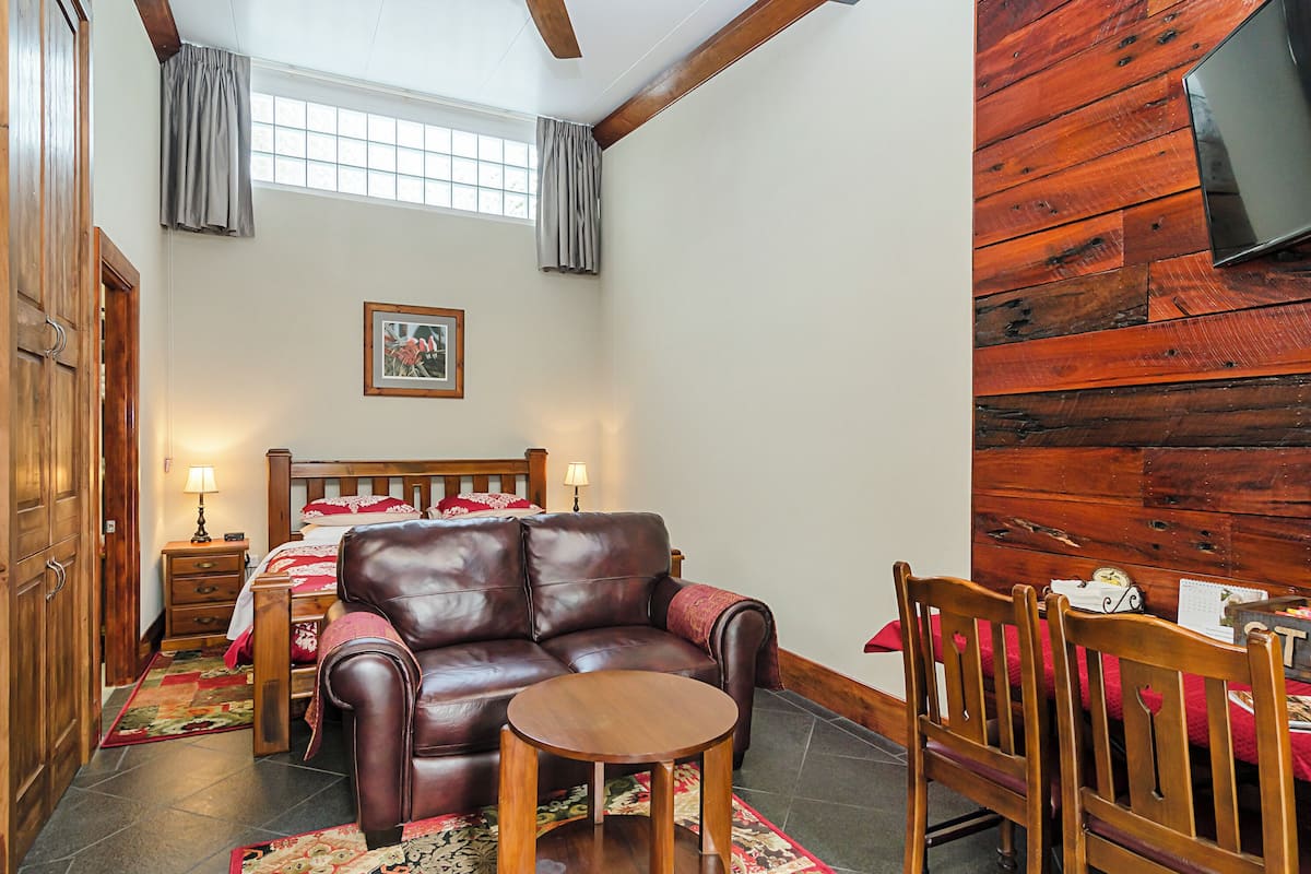 Quarto aconchegante com sofá de couro, uma pequena mesa de madeira e uma cadeira de madeira. Há uma cama com colcha vermelha e branca, uma mesa de cabeceira com abajur e uma janela com cortinas.
