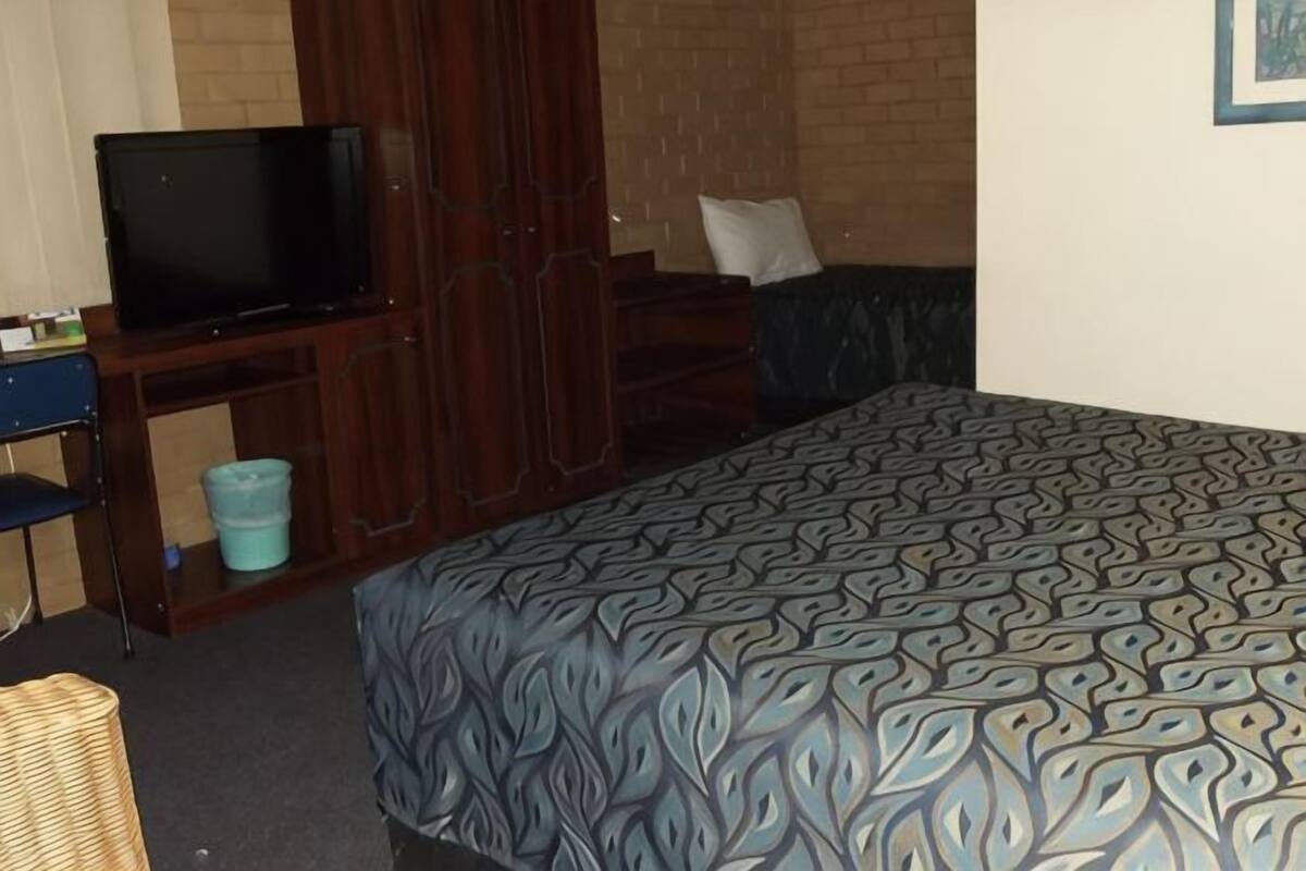 Quarto com uma cama, televisão sobre suporte, uma cadeira e uma mesinha.