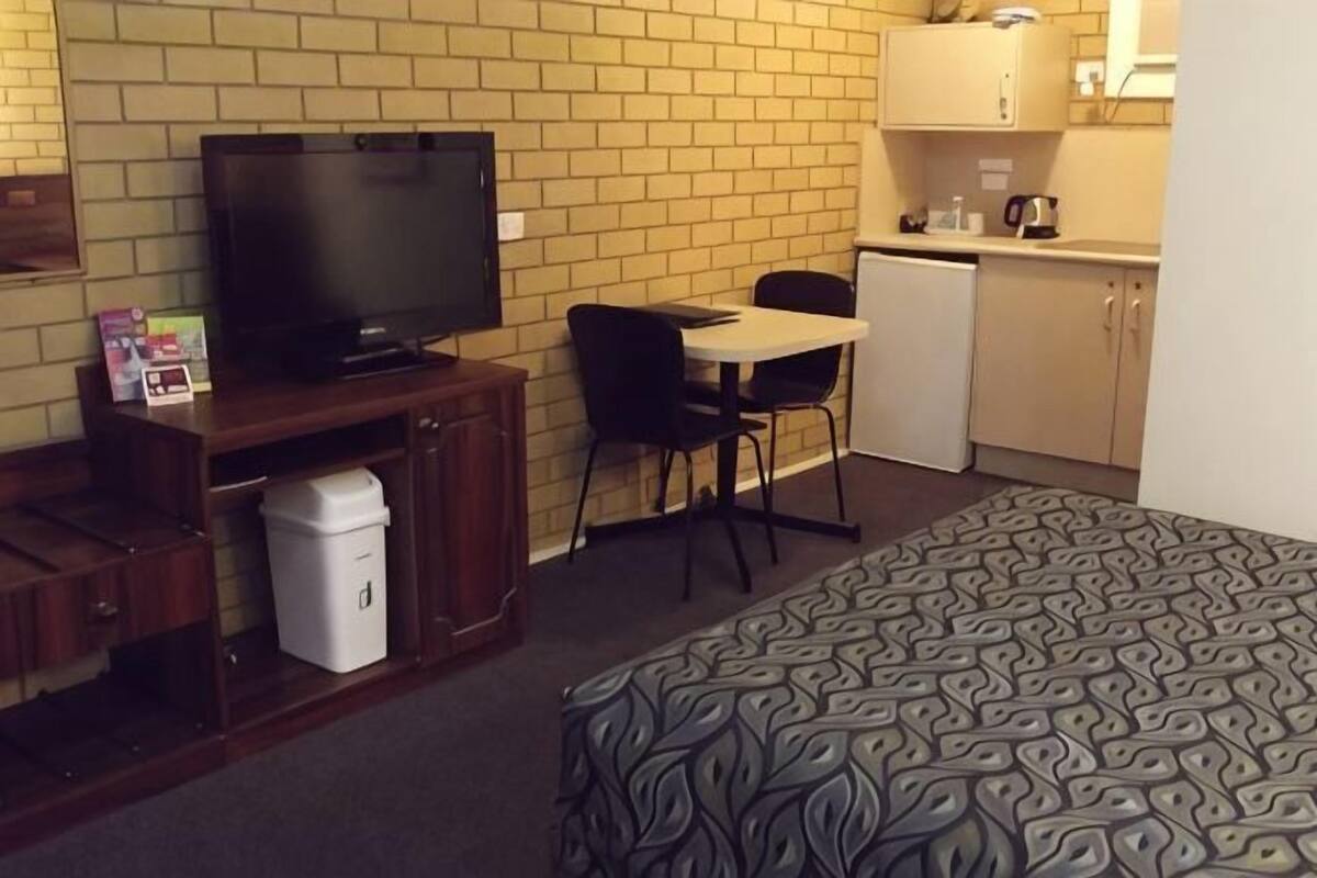 Quarto de hotel compacto com cama, televisão, pequena escrivaninha e cozinha compacta.